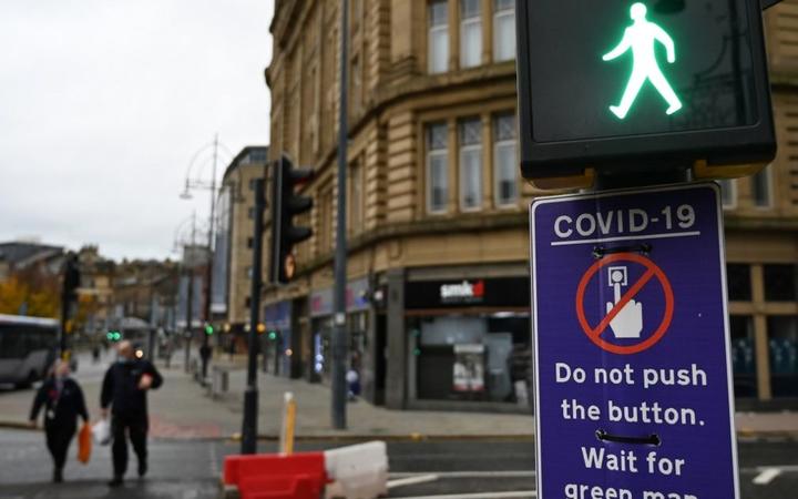 Pedestrians pass a sign displaying a COVID-19 sign advising pedestrians not to use the button, is displayed on a traffic light pole in the centre of Bradford, west Yorkshire on October 31, 2020, as the number of cases of the novel coronavirus COVID-19 rises. 